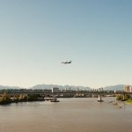 Avião de passageiros sobrevoa um rio diante de uma paisagem urbana e montanhosa.
