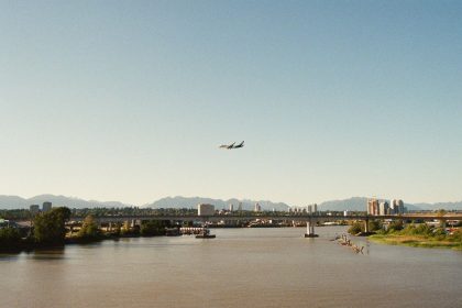 Avião de passageiros sobrevoa um rio diante de uma paisagem urbana e montanhosa.