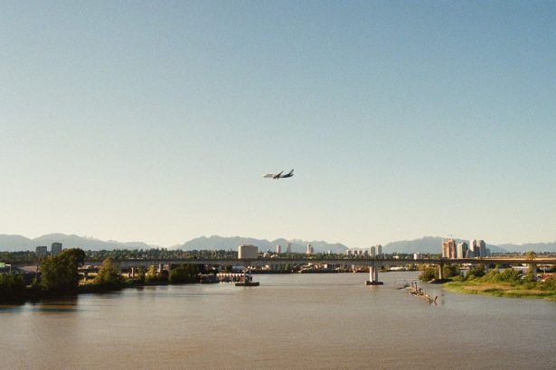 Avião de passageiros sobrevoa um rio diante de uma paisagem urbana e montanhosa.