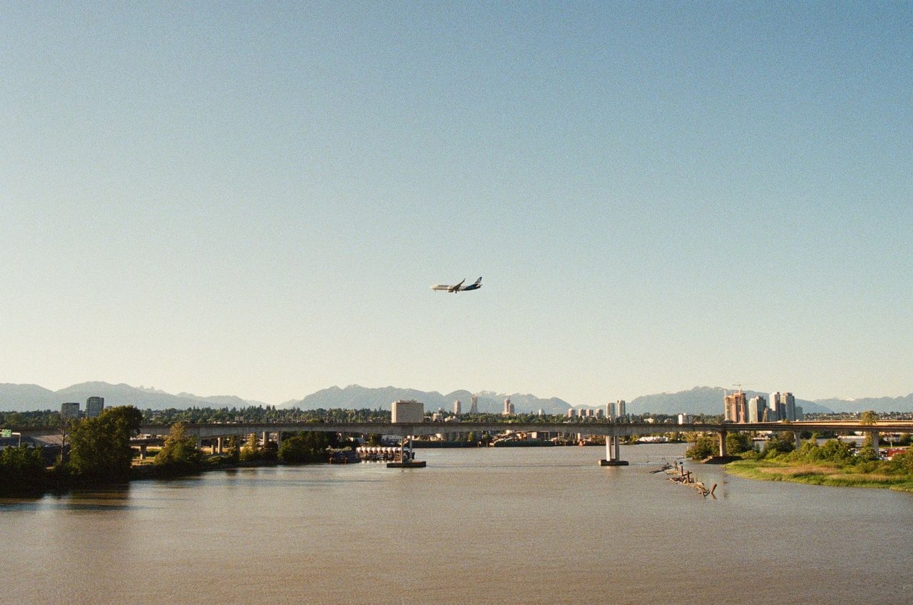 Avião de passageiros sobrevoa um rio diante de uma paisagem urbana e montanhosa.