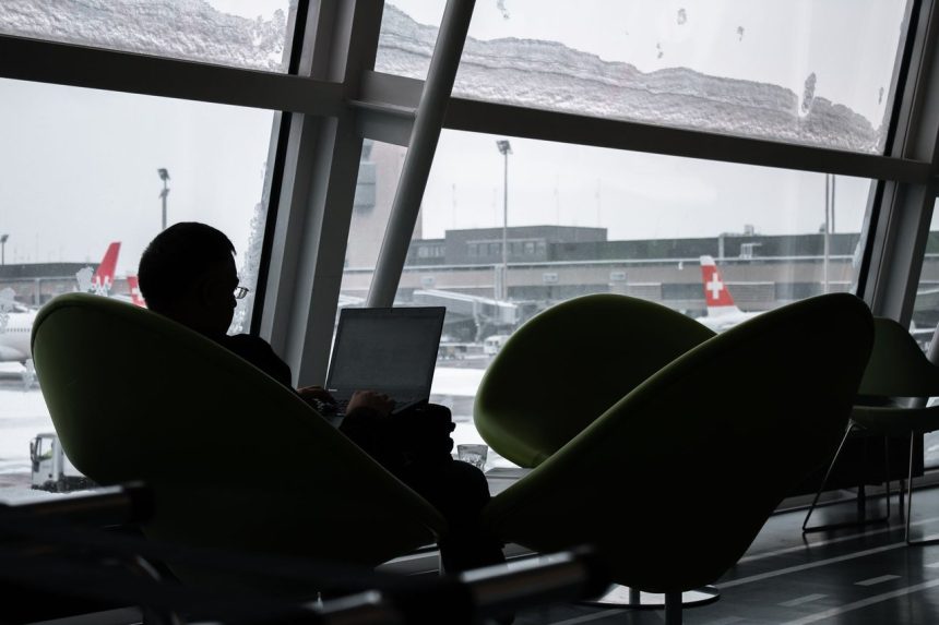 Pessoa usando um laptop no saguão de um aeroporto, com aviões visíveis ao fundo.